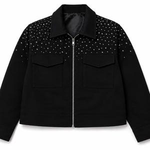 Puede incluir: Una chaqueta negra con cremallera frontal plateada, cuello clásico y dos bolsillos en el pecho con solapa. La parte superior del pecho y los hombros están adornados con numerosas tachuelas plateadas pequeñas y dispersas en un efecto degradado.