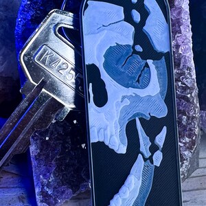 May include: A rectangular black keychain tag featuring a white, detailed graphic of a human skull profile. The tag is attached to a silver metal key with the code K4255 stamped on its surface. The design shows the skull's jaw and eye socket.
