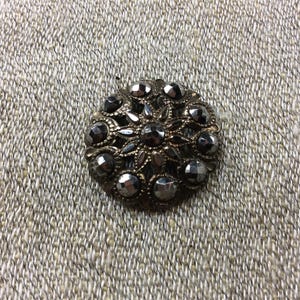 May include: A vintage, circular brooch with a dark, metallic finish. The brooch features a central floral design surrounded by faceted, gray-toned stones. The intricate details and aged appearance suggest a classic, antique style.