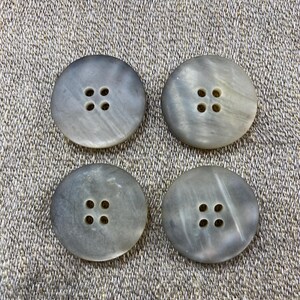 May include: Four round, gray buttons with four holes each, arranged in a square pattern. The buttons have a marbled appearance and are set against a textured, neutral-toned fabric.