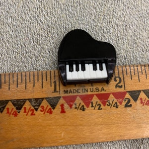 May include: A small black plastic toy grand piano with white keys. The piano is about 1 inch long and is sitting next to a ruler.