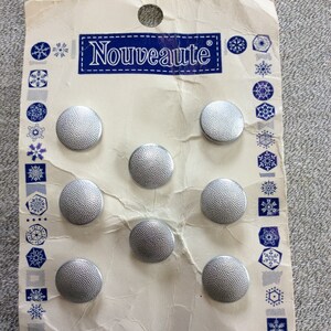 May include: A card with the word "Nouveauté" printed on it. The card has seven silver buttons with a textured surface.