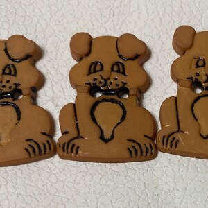 May include: Three brown plastic buttons shaped like dogs with black painted details. The buttons have two holes for sewing.