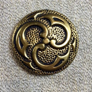 May include: A round, antique brass button with an ornate, swirling design. The button features a textured background and a raised, three-part floral pattern. The button is set against a neutral, textured fabric.