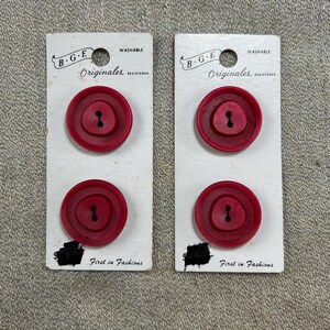 May include: Two sets of vintage red buttons on white cards. Each card has two buttons, with the text "B.G.E Originales" and "First in Fashions". The buttons are a deep red color and have a two-hole design.