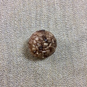 May include: A round, brown decorative object with a mosaic-like surface. The object is composed of small, rectangular pieces in varying shades of brown and amber. The background is a textured, neutral-toned fabric.