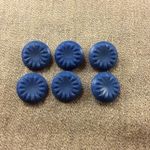 May include: Six navy blue buttons with a carved floral design. The buttons are round and have a slightly raised center. They are arranged in two rows of three on a textured, tan fabric.