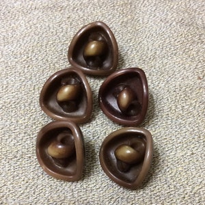 May include: Five brown, triangular buttons with a lighter brown, rounded center. The buttons have a glossy finish and are arranged on a textured, tan surface. These buttons are suitable for sewing and craft projects.