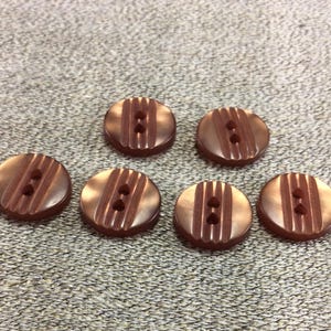 May include: Six brown, round buttons with two holes each. The buttons have a ridged, vertical design and a glossy finish. They are arranged on a textured, light brown surface.