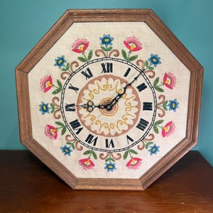 May include: An octagonal wall clock with a wooden frame and a cream-colored fabric face. The face features hand-embroidered pink and blue flowers in a circular pattern, black Roman numerals, and ornate black metal clock hands.