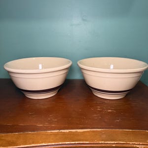Puede incluir: Un par de cuencos de cerámica de color blanco roto a juego, con una banda horizontal de color marrón oscuro que rodea la parte inferior exterior. Cada cuenco tiene un borde ancho y redondeado y un cuerpo curvo que se estrecha hacia una base circular.