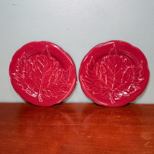 May include: Two round, glossy red ceramic plates featuring a raised, embossed maple leaf design in the centre. Each plate has a scalloped, wavy edge and a smooth, reflective finish, showcasing the detailed veins and texture of the leaf motif.