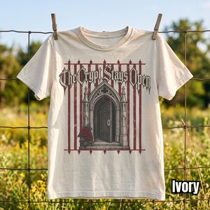 Pode incluir: Uma t-shirt cor de marfim com uma estampa gráfica de um arco de pedra gótico com uma porta aberta, rosas vermelhas e listras verticais vermelhas. O texto "The Crypt Stays Open" está impresso numa fonte gótica estilizada sobre a imagem.
