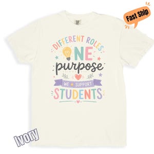 May include: An ivory short-sleeve t-shirt featuring a colourful graphic with the text "DIFFERENT ROLES ONE PURPOSE WE SUPPORT STUDENTS" alongside a lightbulb icon, a heart, and small stars. The design uses pastel pink, yellow, blue, and purple.
