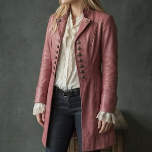 May include: A dusty rose leather coat featuring a high collar, long sleeves with cuffs, and two vertical rows of dark metallic buttons down the front. The coat is worn over a white button-down shirt with ruffled trim and dark blue denim jeans.