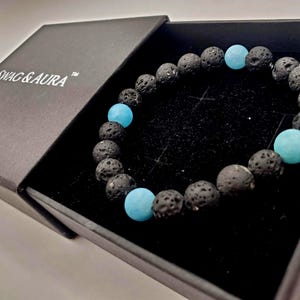 Puede incluir: Una pulsera de cuentas con piedras de lava negras porosas intercaladas con cuentas redondas y lisas de color azul claro. La pulsera se presenta dentro de una caja negra con el texto SWAG & AURA impreso en blanco en la tapa del recipiente.