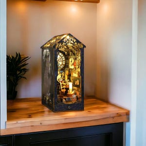Può includere: Un diorama in miniatura a forma di angolo lettura, caratterizzato da una struttura scura e ornata con tetto a capanna. L'interno è illuminato e rivela uno studio dettagliato con orologio, scrivania, sedia e vari piccoli oggetti.