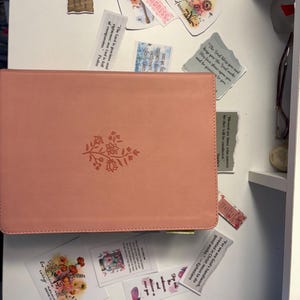 Op de afbeelding: Een rechthoekig boek met een kaft van oudroze kunstleer en een klein, in reliëf aangebracht bloemmotief op de voorkant. Rondom het boek liggen diverse rechthoekige en gestanste stickers met bloemillustraties en religieuze teksten.