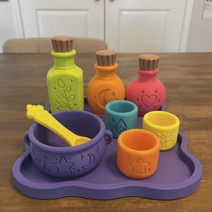May include: A set of colorful silicone potion-making toys including a purple tray, a purple cauldron with a yellow spoon, three bottles with brown stoppers, and three small cups. The items feature embossed icons like leaves, moons, hearts, and stars.