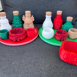 May include: A collection of holiday-themed items including two round trays in red and green, various small bottles shaped like a snowman, Christmas tree, reindeer, and Santa, along with small bowls, a mug, and a red square container with a bow.
