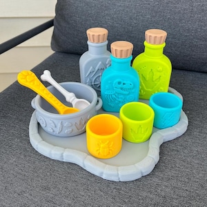May include: A set of silicone children's play items including a gray tray, a gray bowl with a white bone-shaped spoon, two bottles in light blue and lime green, and three small cups in yellow, lime green, and light blue, all featuring embossed designs.