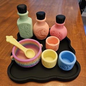 May include: A set of three pastel-colored bottles with black caps, a rainbow-colored cauldron with a matching spoon, and three small cups in peach, yellow, and blue, all arranged on a black tray. Each item features embossed icons.