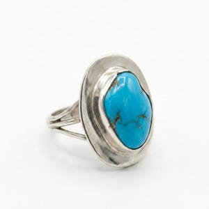 May include: A silver ring featuring a large, oval-shaped turquoise stone with dark brown matrix veins. The stone is set in a smooth, polished silver bezel surrounded by a wide, flat, circular silver plate. The band consists of two split silver wires.