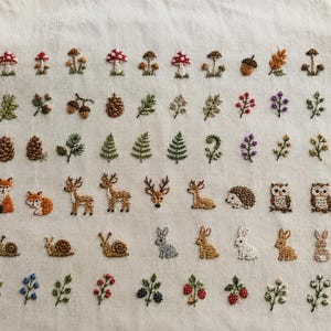 May include: A cream-colored fabric featuring a grid of small, detailed embroidery motifs including various mushrooms, pinecones, acorns, ferns, leaves, berries, and woodland animals such as foxes, deer, hedgehogs, owls, and rabbits.