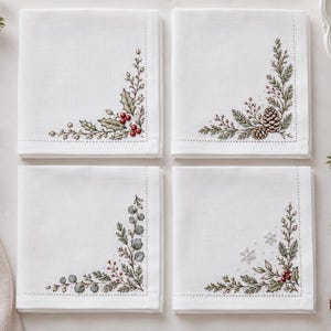May include: Four square white cloth napkins, each featuring a unique embroidered corner design. The patterns include holly leaves with red berries, pine branches with pinecones, eucalyptus sprigs, and winter greenery with small snowflakes.