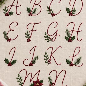 Könnte beinhalten: Eine Sammlung gestickter Buchstaben von A bis N in einer dunkelroten Schreibschrift auf hellbeigem Stoff. Jeder Buchstabe ist mit grünen Stechpalmenblättern und kleinen roten Beeren verziert, außer dem Buchstaben M, der einen roten Weihnachtsstern zeigt.
