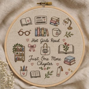 Può includere: Un telaio da ricamo circolare in legno contenente tessuto beige con disegni ricamati a mano di libri, una tessera della biblioteca, occhiali, una tazza, una lampada, una borsa e piante. Il testo recita Hot Girls Read e Just One More Chapter.