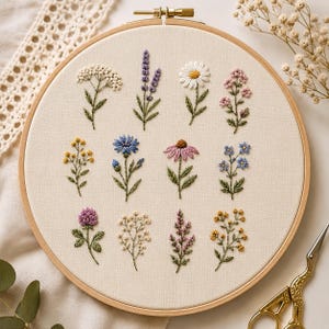 Puede incluir: Un bastidor de bordado circular de madera que contiene tela de color blanco roto con doce diseños florales cosidos a mano. Las flores incluyen lavanda, margaritas, acianos y tréboles en tonos morados, azules, amarillos, rosas y blancos.
