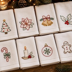May include: A set of eight white cloth napkins, each featuring a distinct embroidered Christmas motif including a tree, poinsettia, bells, holly, candy cane, candle, ornament, and gingerbread person, all with a hemstitched border detail.