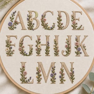 Puede incluir: Un bastidor de bordado circular de madera que contiene tela de color blanco roto con las letras de la A a la N bordadas en hilo color canela. Cada letra está decorada con pequeñas flores bordadas de colores y hojas verdes en tonos rosa, azul y amarillo.