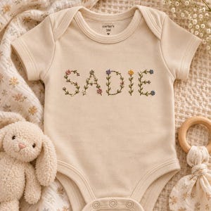 Peut inclure: Un body pour bébé à manches courtes de couleur crème avec le prénom SADIE brodé dans une police de caractères en forme de vigne florale, orné de petites fleurs roses, violettes, jaunes et bleues, un col rond et trois boutons-pression.