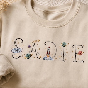 Könnte beinhalten: Ein beigefarbenes Sweatshirt mit Rundhalsausschnitt und dem gestickten Namen SADIE in schwarzen Buchstaben. Jeder Buchstabe ist mit Weltraummotiven verziert, darunter eine Spiralgalaxie, Planeten mit Ringen, eine startende Rakete und kleine goldene Sterne.