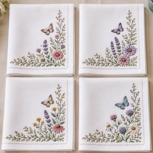 Könnte beinhalten: Ein Set aus vier quadratischen weißen Servietten, jede mit einer einzigartigen Eckstickerei aus bunten Wildblumen und einem einzelnen Schmetterling. Die Blumenmotive umfassen Rosa-, Lila-, Gelb- und Grüntöne mit feinen gestickten Details.