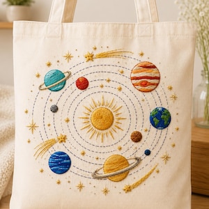 May include: A cream-colored canvas tote bag featuring an intricate hand-embroidered solar system design. The center displays a bright yellow sun surrounded by concentric dashed orbits, various planets in shades of blue, orange, and brown, and gold stars.