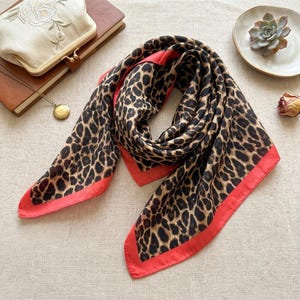 Puede incluir: Un pañuelo cuadrado con un estampado de leopardo en marrón y negro con un borde rojo coral sólido. La tela está dispuesta en una forma suelta y plegada, resaltando el contraste entre el estampado animal y los bordes rojos vibrantes.