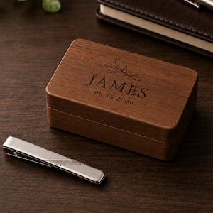 Puede incluir: Una caja rectangular de madera marrón oscuro con tapa abatible, grabada con el nombre JAMES y la fecha 15.06.2026 bajo una pequeña ilustración de hoja. Un pasador de corbata de metal plateado con un patrón de líneas diagonales.