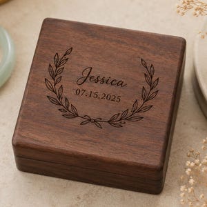 May include: A square dark brown wooden jewelry box featuring a laser-engraved laurel wreath design on the lid. The center of the wreath displays the name Jessica and the date 07.15.2025 in a script and sans-serif font combination.