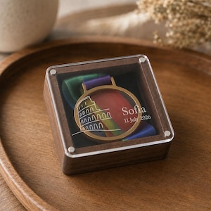 May include: A square wooden box with a clear acrylic lid containing a gold-toned circular medal. The lid features a white line drawing of the Colosseum, the name Sofia, and the date 11 July 2026. A multi-colored ribbon is visible inside the medal.