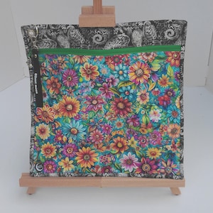 May include: A square fabric pouch featuring a vibrant floral pattern with red, orange, yellow, and blue flowers on a teal background. The pouch has a black and white paisley border, a bright green zipper, and a black zipper pull with white text.