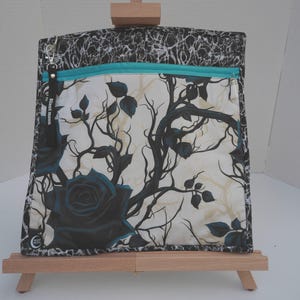 May include: A square fabric pouch featuring a cream background with a black rose and branch pattern. It has a teal zipper closure across the top and a black border with a white marbled design. A black fabric tag with the text Silent Moon is attached.