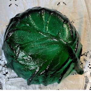 Pode incluir: Uma tigela de vidro verde esmeralda profundo com uma superfície texturizada e moldada que imita a aparência em camadas e nervurada de uma folha de couve. A tigela tem uma forma arredondada e orgânica com um acabamento brilhante.