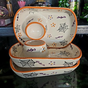 May include: A stack of three rectangular ceramic serving dishes with rounded corners and orange rims. The cream-colored surface features a hand-painted pattern of black spiderwebs, purple bats, and small orange and purple flower clusters.
