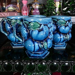 May include: Three matching ceramic mugs featuring a raised, three-dimensional fruit motif in shades of deep blue and green. Each mug has a glossy finish, a sturdy handle, and a hexagonal base, showcasing detailed relief carvings of rounded fruits.
