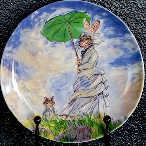 May include: A round decorative plate featuring a painted illustration of an anthropomorphic rabbit wearing a white dress and bonnet, holding a green umbrella. A smaller rabbit stands nearby in a field of green grass and purple flowers under a blue sky.