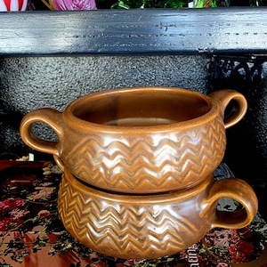 May include: Two stacked brown ceramic bowls, each featuring two side handles and a textured chevron pattern wrapping around the exterior. The bowls have a glossy finish with darker brown accents within the grooves of the zigzag design.