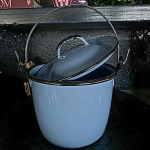 Könnte beinhalten: Ein hellblauer Eimer aus Emaille mit passendem Deckel und einem dunklen Metallgriff. Der Eimer hat eine glatte, glänzende Oberfläche mit einem dunklen Rand an der Oberkante und am Deckel. Der Deckel liegt schräg auf und bedeckt die Öffnung.